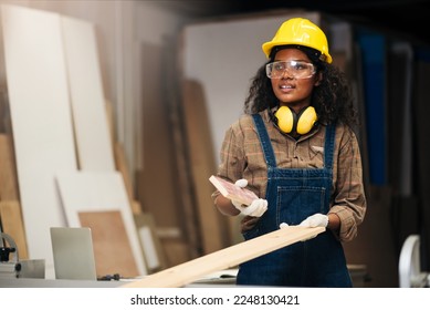 Young attractive black female carpenter wearing safety goggles and hard hat for woodworking in small business workshop. Wood  industry and furniture industry. - Powered by Shutterstock