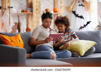 Young Attractive Black Mother Sitting On Sofa In Living Room And Using Tablet While Reading Halloween Story To Son