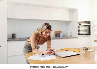 Young attractive blond woman leaning on kitchen table and using smart phone for e-banking. - Shutterstock ID 1823625659