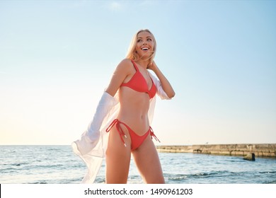Young attractive blond woman in red swimsuit and white shirt happily standing on beach with sea on background