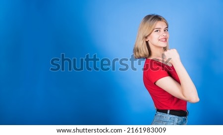 Young and attractive blonde caucasian girl in casual clothes pointing her thumb at copy space for advertising isolated on blue studio background.
