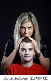 A Young Attractive Blonde Mother Stands Behind Her White Teenager Son And Hugs Him. Studio Photo On A Dark Background