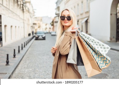 Young attractive blonde woman in autumn coat and sunglasses holding lot of shopping packages and walking along European street. Travel and shopping concept