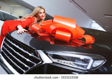 A Young Attractive Blonde Woman In A Red Suit Is Hugging A Large Black Crossover Car With A Huge Red Bow On The Background Of A Car Dealership Or Auto Showroom.