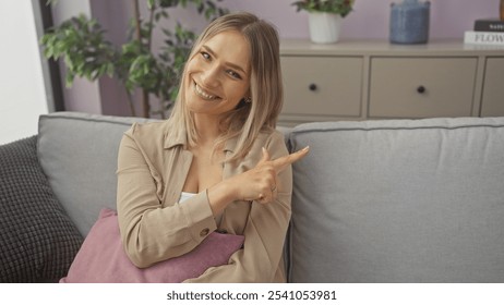 A young, attractive, blonde woman smiling and pointing while sitting indoors in a cozy living room with potted plants and a comfortable couch. - Powered by Shutterstock