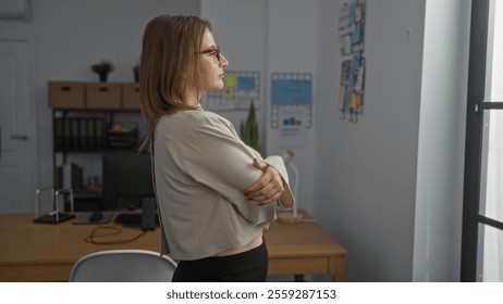 A young, attractive, blonde woman standing with arms crossed in a modern office, wearing professional attire and glasses, looking thoughtfully out the window at her workplace. - Powered by Shutterstock