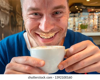 Young Attractive Blue-eyed Man In A Blue Sweater Drinking Cappuccino And Smiling With Happiness With Coffee Foam On His Lips