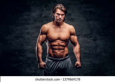Young attractive bodybuilder with naked torso is posing for photographer on grey background.