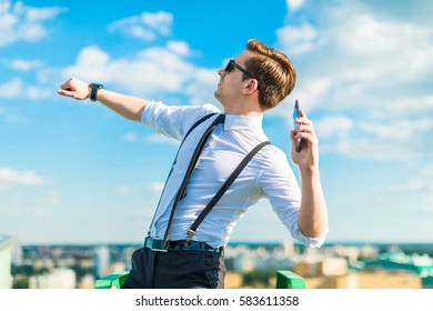 Young Attractive Brunette Caucasian Businessman In White Shirt, Black Tie, Black Braces, Costly Watch And Black Sunglasses Stand On The Green Ladder On The Roof, Throw Phone Out