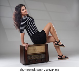 Young Attractive Brunette Caucasian Woman Sitting On A Vintage Radio, Studio Shot On Gray Background With Window Projection