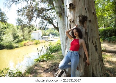 Young, Attractive, Brunette Woman With Curly Hair, Red T-shirt And Jeans, Leaning Against A Big Tree In An Outdoor Park With Provocative Look. Concept Sensuality, Provocation, Beauty, Nature.