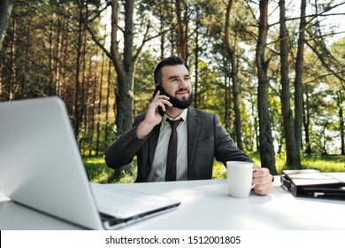 Young attractive business man in suit and tie sits at desk and works on computer outdoors. Drink coffee from white cup and talks phone. Green trees, nature and park on background. Remote work outside - Powered by Shutterstock