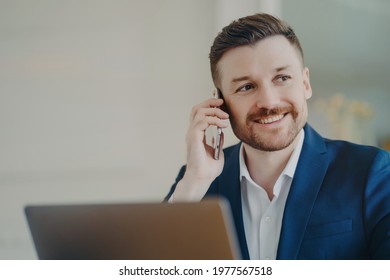 Young attractive businessman in formal suit talking on phone and smiling when hearing good news. Happy manager sitting in office in front of laptop and thinking of positive outcomes of conversation - Shutterstock ID 1977567518