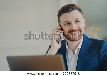 Young attractive businessman in formal suit talking on phone and smiling when hearing good news. Happy manager sitting in office in front of laptop and thinking of positive outcomes of conversation
