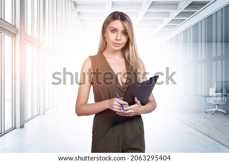 Young attractive businesswoman wearing formal jumpsuit is holding clipboard. Office workplace with panoramic window and concrete floor in the background. Concept of successful career and leadership
