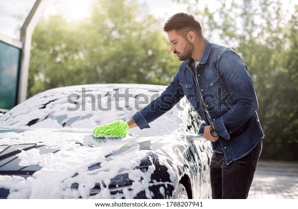 Young attractive Caucasian bearded\
man washing his electric luxury car in a self-service car wash\
station outdoors with cleaning foam and green sponge\
mitten