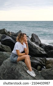 A young attractive Caucasian blonde woman of 35 years sits on the rocks of sea coast. Emerald-colored water, waves splashing on the stones. The woman meditates and relaxes.