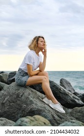 A young attractive Caucasian blonde woman of 35 years sits on the rocks of sea coast. Emerald-colored water, waves splashing on the stones. The woman meditates and relaxes.
