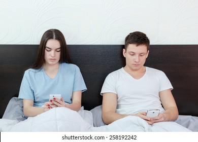 Young attractive caucasian couple with smartphones in pajamas in their bed searching or browsing over internet and texting in chat with other people, cheating over messages, checking each other phones