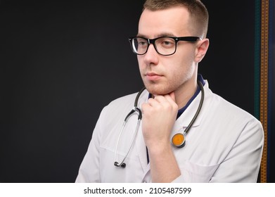 A Young Attractive Caucasian Doctor In Glasses And A White Coat Stands Pensive And Looks To The Side. Studio Portrait Of A Paramedic