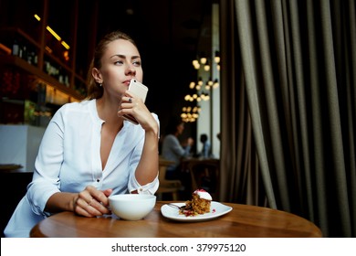 Young attractive Caucasian female holding a mobile phone near face while dreaming about something, charming woman waiting for a call on her cell telephone while sitting in modern restaurant interior 
