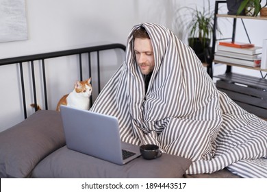 Young attractive caucasian man sitting on a bed wrapped with blanked at home working using laptop with cup of hot drink. Guy browsing Internet for flu, influenza, cold and covid-19 symptoms