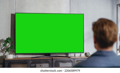 Young Attractive Caucasian Man Sitting On A Sofa And Watching TV With Horizontal Green Screen Mockup. It's Day Time On Weekend At Home. Spacious Living Room In Stylish Loft Apartment.