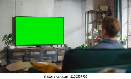 Young Attractive Caucasian Man Sitting On A Sofa And Watching TV With Horizontal Green Screen Mock Up. It's Day Time On Weekend At Home. Spacious Living Room In Stylish Loft Apartment.