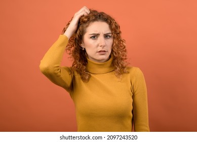 A young and attractive Caucasian redhead girl in an orange jumper holding her hand on her head because she is confused isolated on an orange studio background.