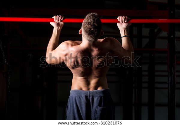 Young attractive caucasian sportsman working out in\
gym, exercising on chin-up bar, doing pull-ups for arms and back\
muscles, rear view