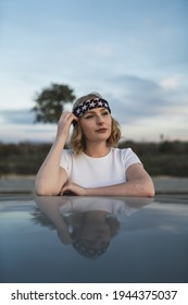 A Young Attractive Caucasian Woman With Blond Hair Wearing A Bandana With A US Flag Leaning On A Car