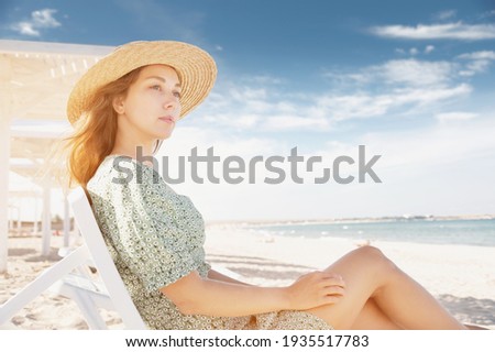 Young attractive caucasian woman in a green dress and a straw sits resting on a lounger under a wooden umbrella on a sandy beach of the seashore or ocean.