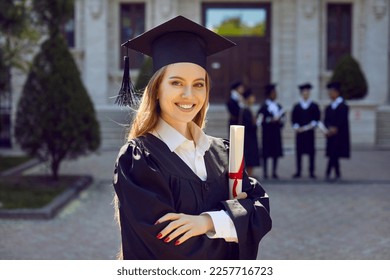 Young attractive Caucasian woman posing smilingly for student portrait holding certificate of higher education grant wearing black academic gown and hat standing in front of college building - Powered by Shutterstock