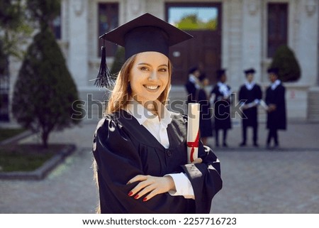 Young attractive Caucasian woman posing smilingly for student portrait holding certificate of higher education grant wearing black academic gown and hat standing in front of college building