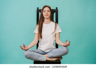 Young Attractive Caucasian Woman Sitting On Chair In Yoga Meditation Pose With Calm Face, Looking At Camera, Dressed In Casual Over Turquoise Studio Background. Mockup, Balance, Confidence. Copy Space