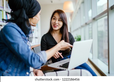 A young and attractive Chinese Asian woman has a business meeting with a Malay Muslim woman. They are both sitting in an office and talking over a laptop computer during the day. 