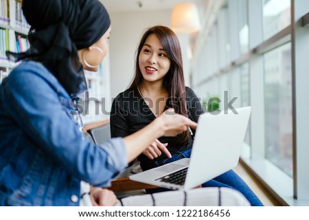 A young and attractive Chinese Asian woman has a business meeting with a Malay Muslim woman. They are both sitting in an office and talking over a laptop computer during the day. 