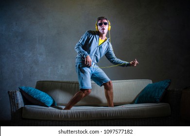 Young Attractive And Cool Happy Man Playing Air Guitar Listening To Music With Yellow Headphones Holding Mobile Phone Jumped On Top Of Home Sofa Couch Excited In Trance Isolated On Grunge Bg