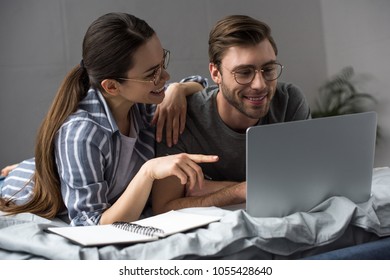 Young attractive couple with notepad looking at laptop while lying in bed