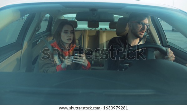 Young attractive\
couple sitting in auto, starting car. Girl with smartphone in hands\
using navigator app, showing way to boyfriend. Destination. Strange\
city. Outdoors.