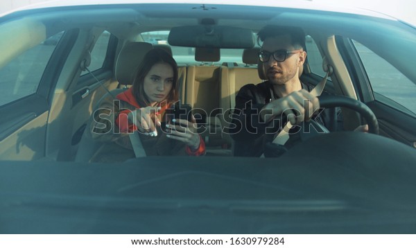 Young attractive\
couple sitting in auto, starting car. Girl with smartphone in hands\
using navigator app, showing way to boyfriend. Destination. Strange\
city. Outdoors.