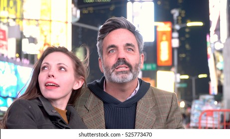 Young Attractive Couple Sitting In Times Square New York City Looking At The Bright Billboard Neon Signage In Cold Weather. Location For New Years Eve Celebration When Ball Drops