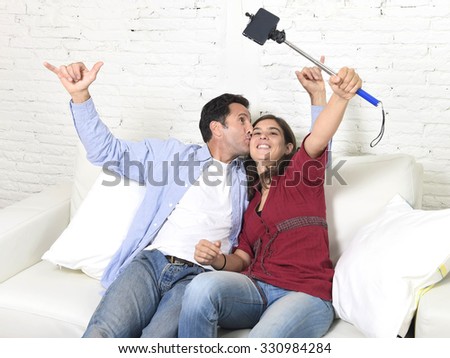 young attractive couple taking selfie photo or shooting self video with mobile phone and stick sitting at home couch smiling happy together man kissing woman celebrating their love