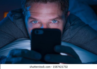 Young Attractive And Crazy Internet Addict Man With Blue Eyes Lying On Bed Late At Night In Dark And Dim Light Networking On Mobile Phone Or Online Dating In Social Media Addiction And Overuse