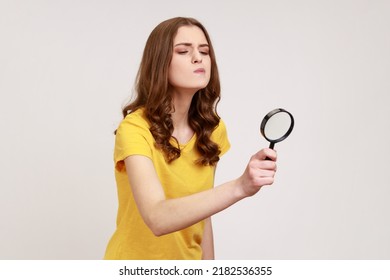 Young Attractive Curious Female With Brown Hair Looking Through Magnifying Glass, Spying, Finding Out Something, Inspecting Crime Scene. Indoor Studio Shot Isolated On Gray Background.