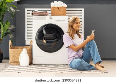 Young attractive cute woman, housekeeper holding smartphone using mobile app sitting on carpet in modern laundry room with washing machine, laundry detergent, basket with clothes on background. Mockup - Powered by Shutterstock