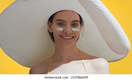 Young Attractive Dark-haired Woman In A Big White Hat Applies Sunblock On Cheeks And Enjoys The Sun Smiling Wide Against Yellow Background | Sunscreen Application Shot For Skincare Commercial