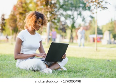 Young Attractive Dark-skinned College Student Wearing Tank Top And Jeans Sitting On The Lawn At Campus On Sunny Day, Working On Her Thesis Using Laptop Computer, Looking Busy And Concentrated