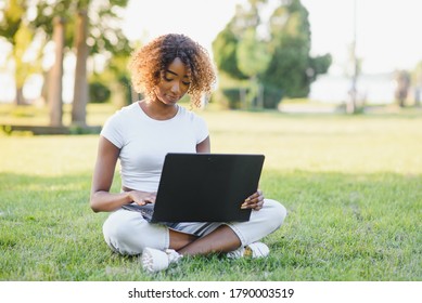 Young Attractive Dark-skinned College Student Wearing Tank Top And Jeans Sitting On The Lawn At Campus On Sunny Day, Working On Her Thesis Using Laptop Computer, Looking Busy And Concentrated