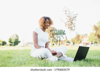 Young Attractive Dark-skinned College Student Wearing Tank Top And Jeans Sitting On The Lawn At Campus On Sunny Day, Working On Her Thesis Using Laptop Computer, Looking Busy And Concentrated